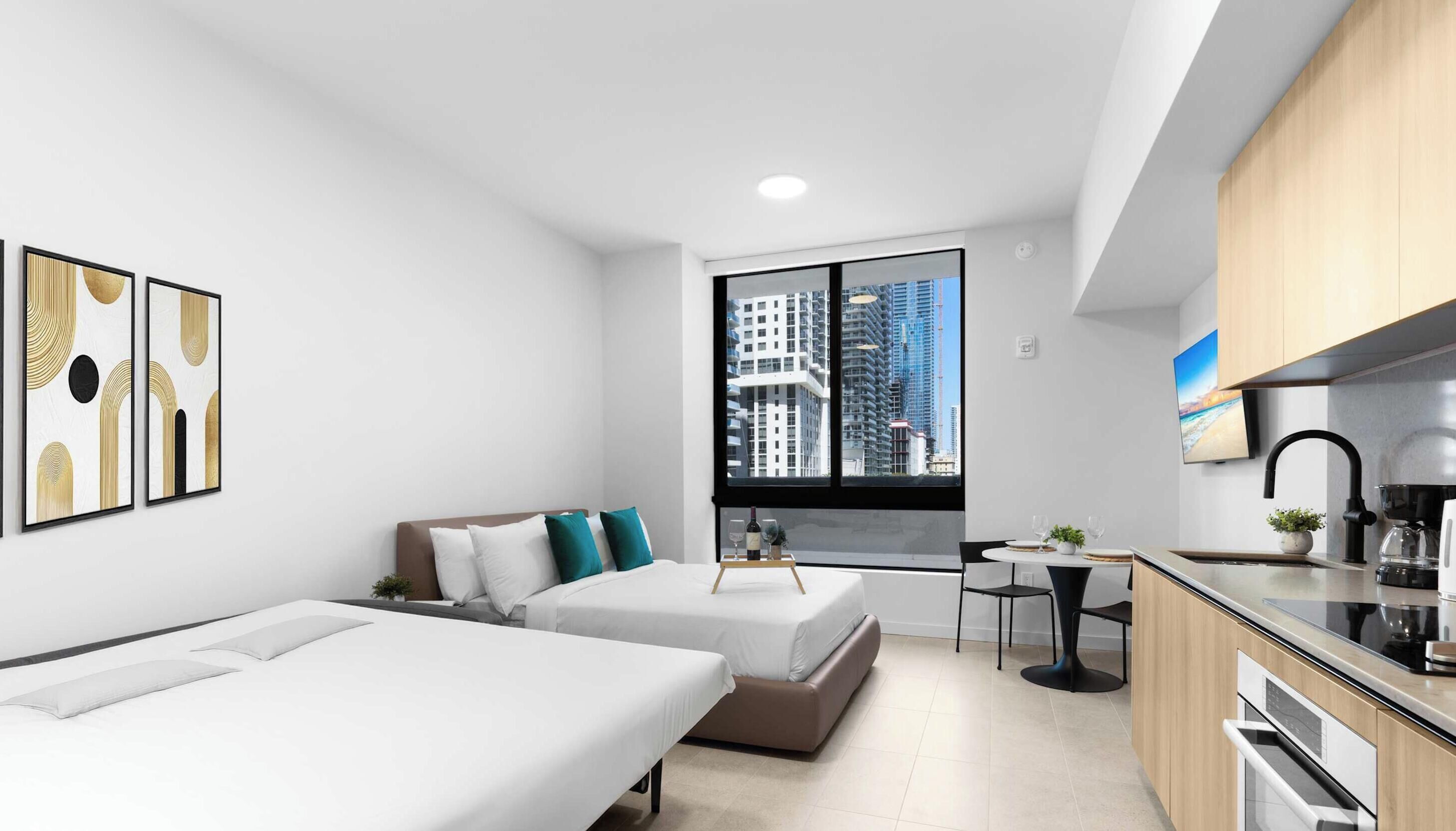 Une chambre d’hôtel moderne avec un lit, une petite table sur laquelle se trouve une bouteille de vin, une vue sur les immeubles, un coin cuisine et une grande fenêtre.