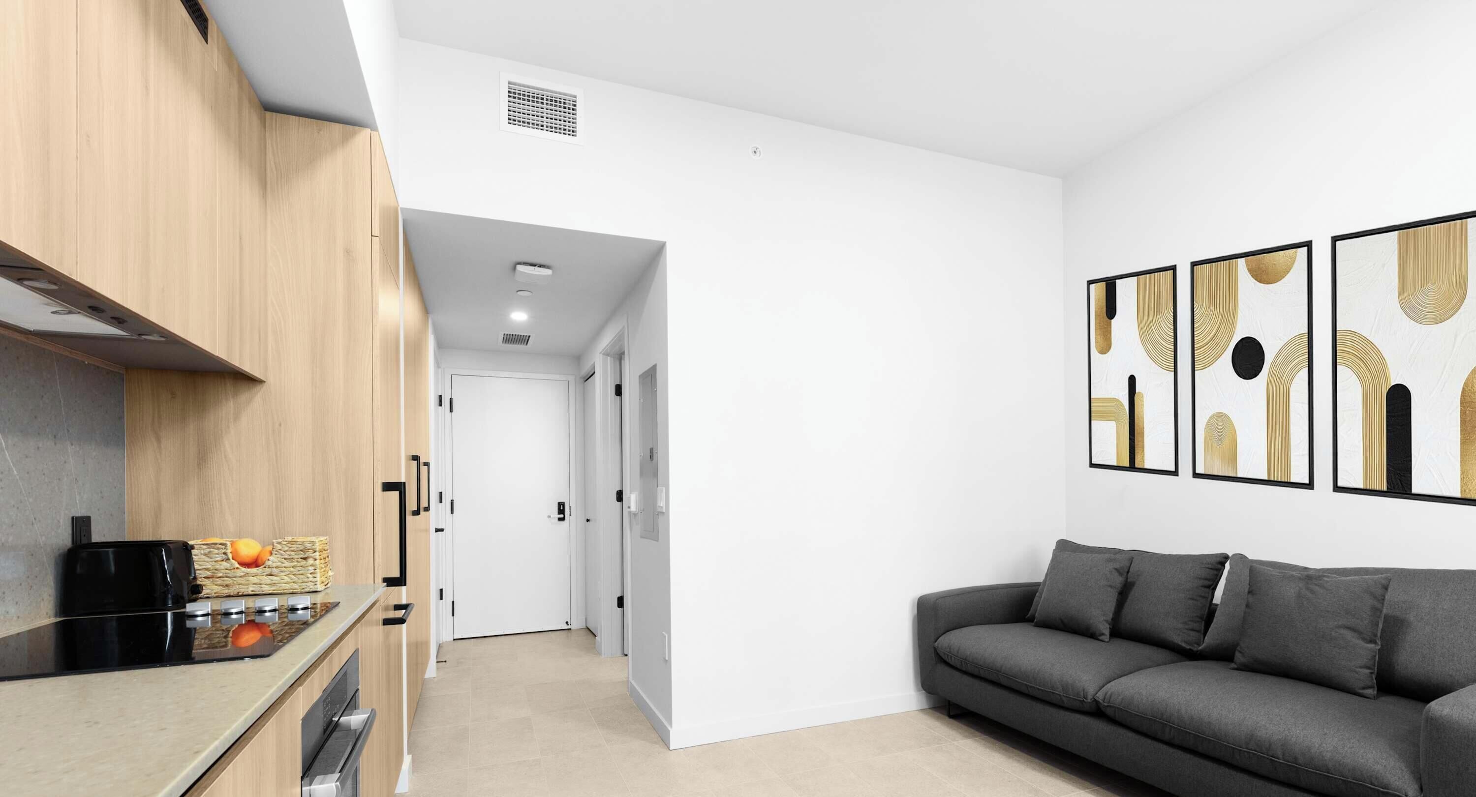 A modern kitchen with a built-in stove, a wooden cabinet, and a countertop. There is a gray sofa and a wall with abstract art.