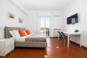 Quarto com cama, escrivaninha, cadeira, televisão e varanda.