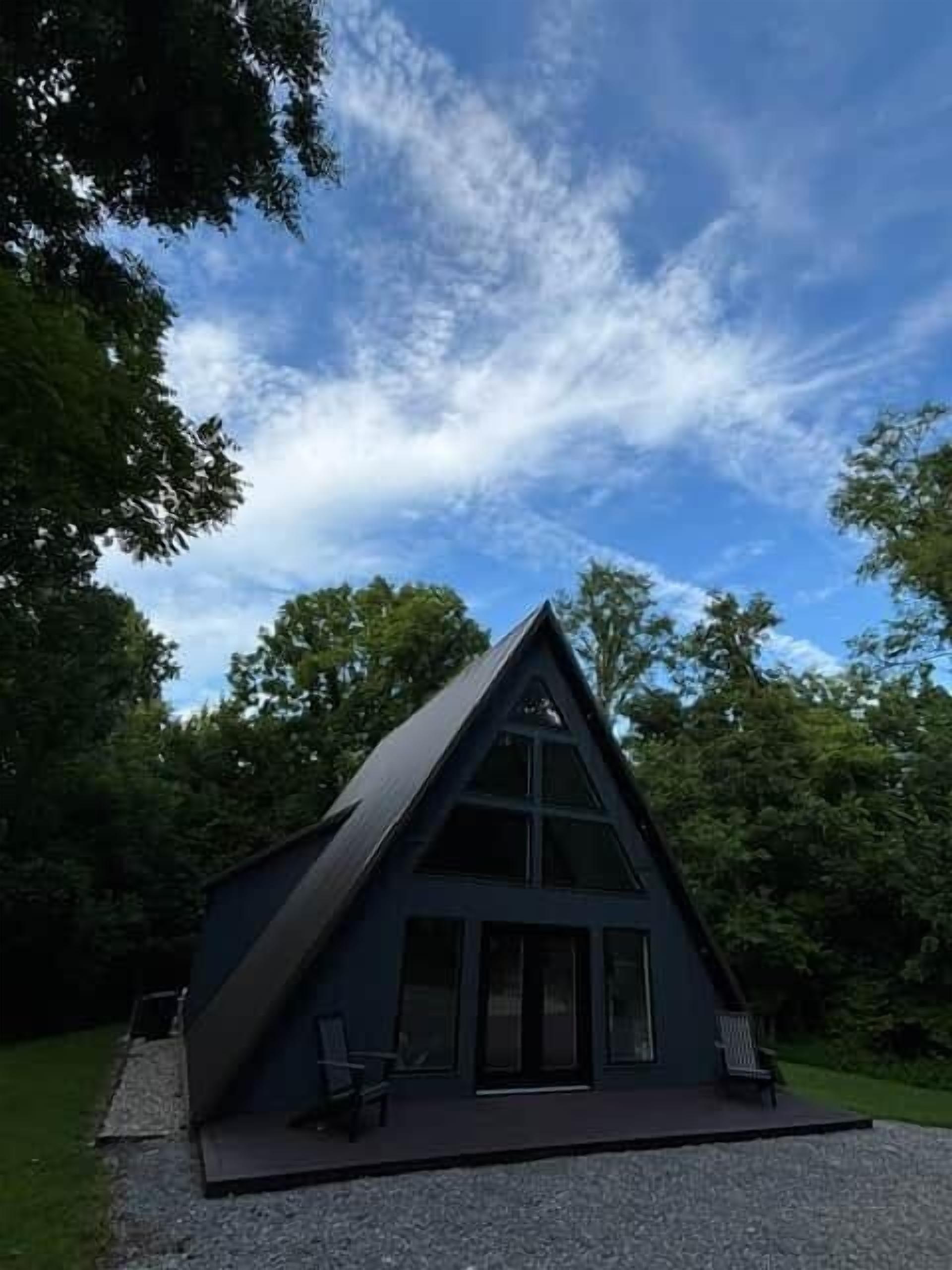 Une cabane moderne aux couleurs sombres, dotée d’un toit triangulaire, de grandes fenêtres et d’une allée gravillonnée.