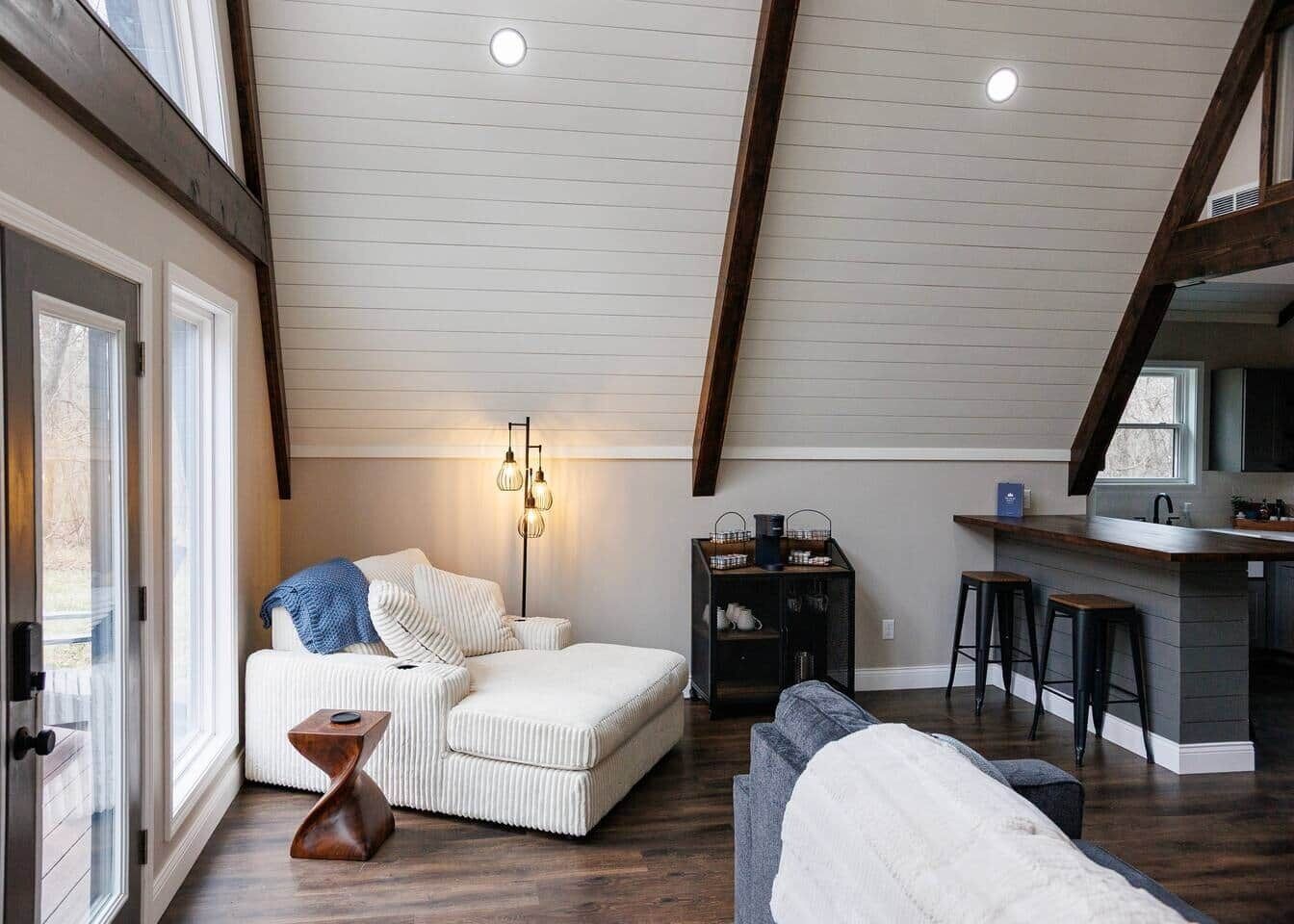 A loft-style room with a high ceiling, a built-in kitchen area, and a seating area with a sofa and ottoman.