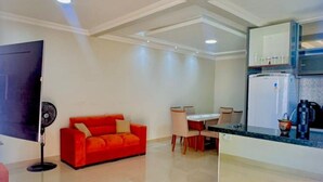 A modern living room with a red sofa, a dining area with a table and chairs, a kitchen with a refrigerator, and a flat-screen TV mounted on the wall.