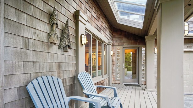 Un'area esterna con una terrazza in legno, sedie Adirondack, un ingresso con porta in vetro e un muro decorativo con un pino.