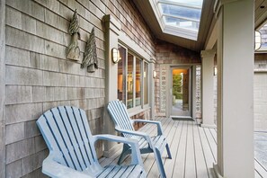 Une terrasse en bois avec des fauteuils Adirondack, une porte d’entrée en verre et un mur décoratif orné d’un pin.
