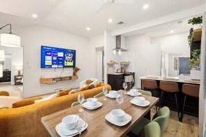 Ein moderner Essbereich mit einem Holztisch, gedeckt für vier Personen, einer Couch und einem Fernseher, der Streaming-Dienste anzeigt.