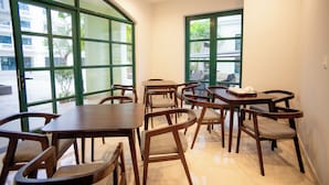 Un'area pranzo con diversi tavoli e sedie, ampie finestre a cornice verde e vista sull'esterno.