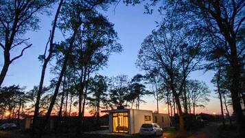 A small, modern house with a glass door, surrounded by tall trees and a sunset in the background.