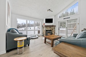 A cozy living room with a stone fireplace, large windows, and a flat-screen TV mounted on the wall.