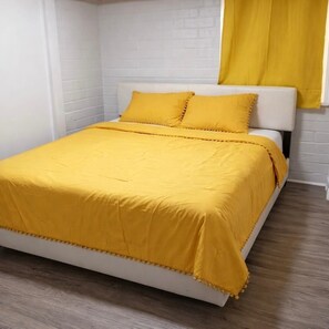 Una cama con colcha y almohadas amarillas, cabecera blanca y suelo de madera.
