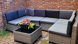 En liten utomhusavdelning med sittplatser i ett hörn, med en soffa i rotting och en blomkruka med blommor.