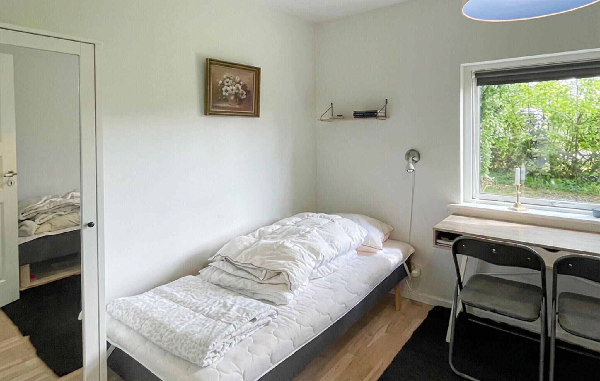 Ein Einzelbett mit weißer Daunendecke, ein Schreibtisch mit Stuhl, ein Fenster mit Blick ins Grüne und ein Wandregal.