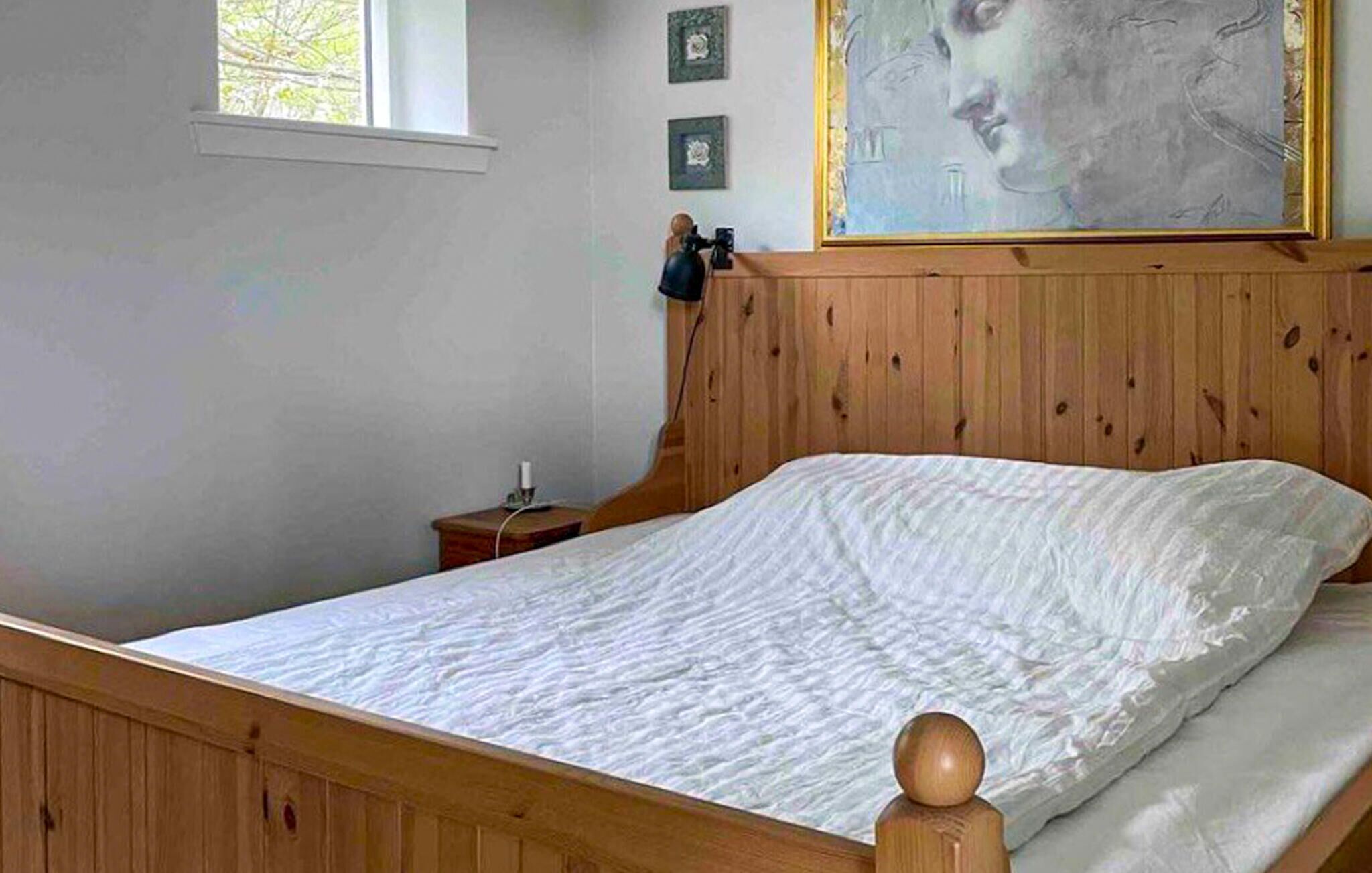 Una cama de madera con colchón blanco en una habitación con cabecera de madera, una mesita de noche pequeña y un cuadro en la pared.