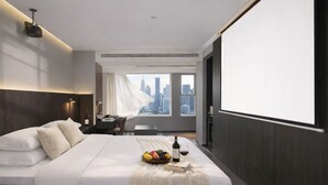 A hotel room with a bed, a desk, a chair, a large window with a city view, and a projector screen. - Chengdu Suli Travel Hotel (Chengdu)
