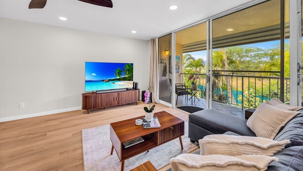 A modern living room with a TV, wooden coffee table, sofa, and a balcony with a view of greenery. -  Kihei Kai Nani Beachside Resort Vistas (Kihei)