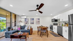 A modern kitchen with white cabinets, a dining area with a wooden table and chairs, a gray sofa, and a ceiling fan. -  Kihei Kai Nani Beachside Resort Vistas (Kihei)