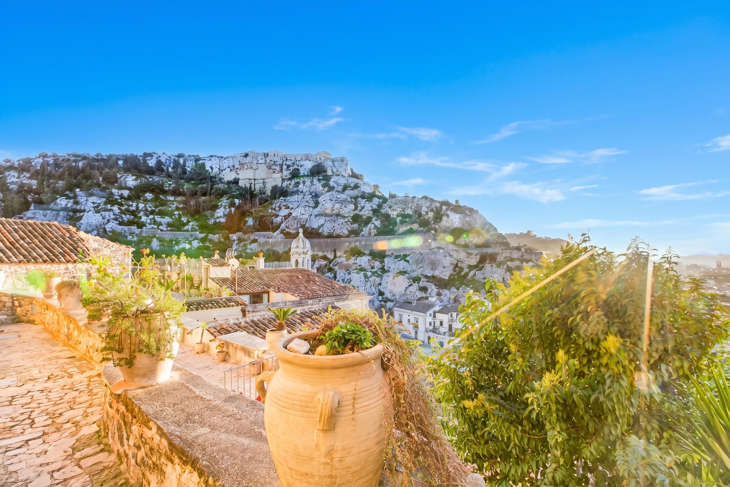 A traditional village with stone buildings, a large terracotta pot, and a view of a hill with ruins.