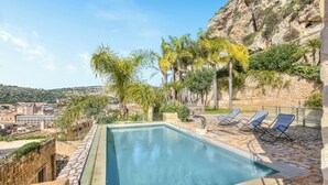 A swimming pool with lounge chairs, a stone wall, and a view of a cityscape.