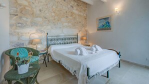 Ein Zimmer mit Steinwand, einem Bett mit weißer Bettwäsche, einem grünen Stuhl und einem Gemälde an der Wand.