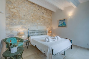Una stanza con parete in pietra, un letto con biancheria bianca, una poltrona verde e un quadro appeso al muro.