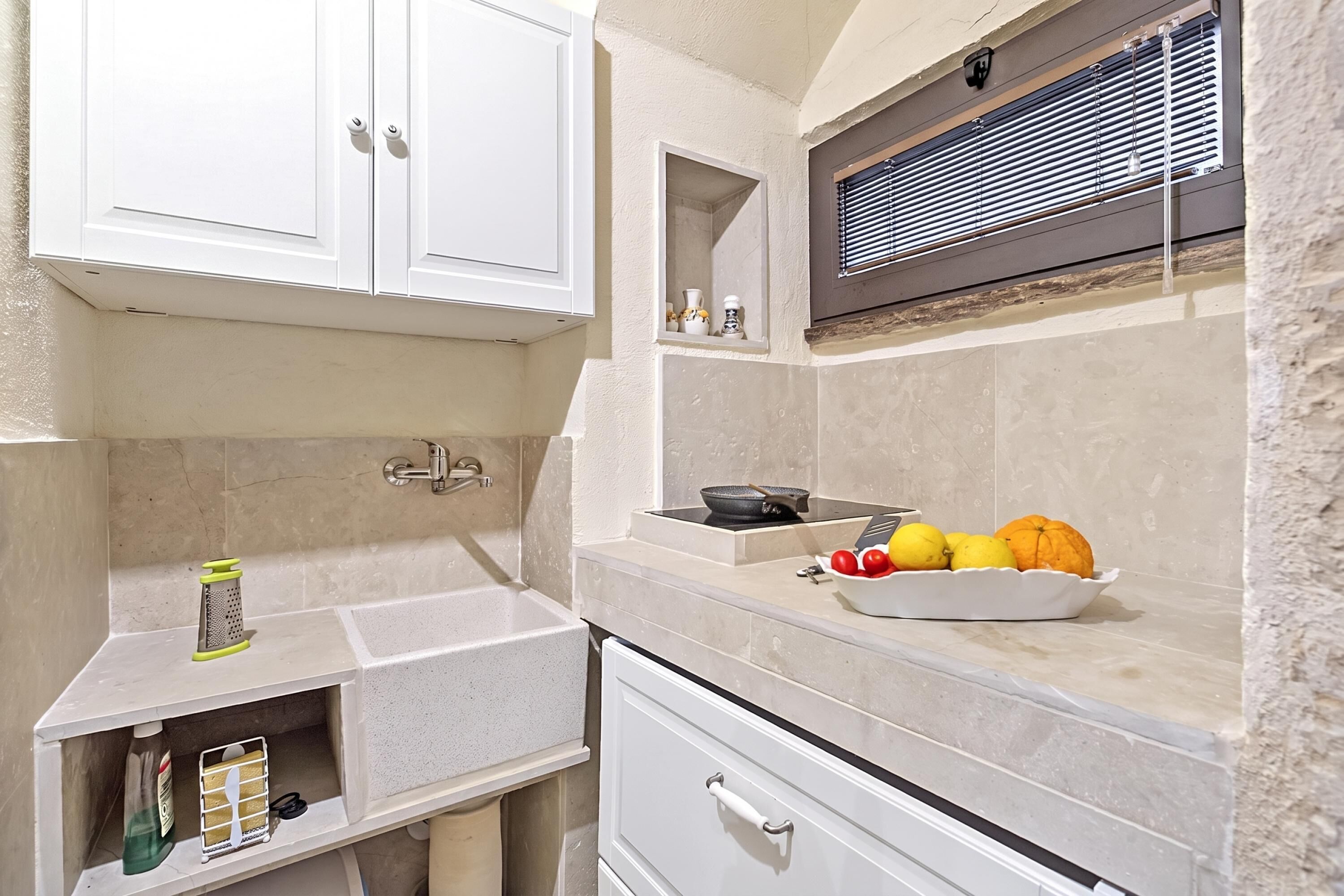 A kitchen with white cabinets, a countertop, a sink, a window with blinds, and a bowl of fruit.