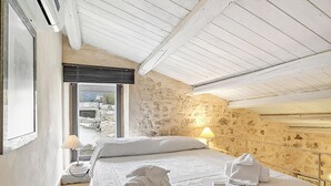 Ein Schlafzimmer mit Steinsandsteinwand, einem Bett mit weißen Leinentüchern, einem Fenster mit Aussicht und einer Lampe auf dem Nachttisch.
