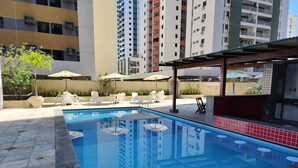 Una piscina con bordes de azulejos, rodeada de sillas de descanso y sombrillas, con edificios residenciales altos al fondo.