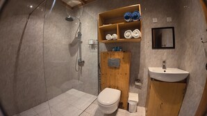 A bathroom with a shower, toilet, wooden shelf with towels, and a sink.