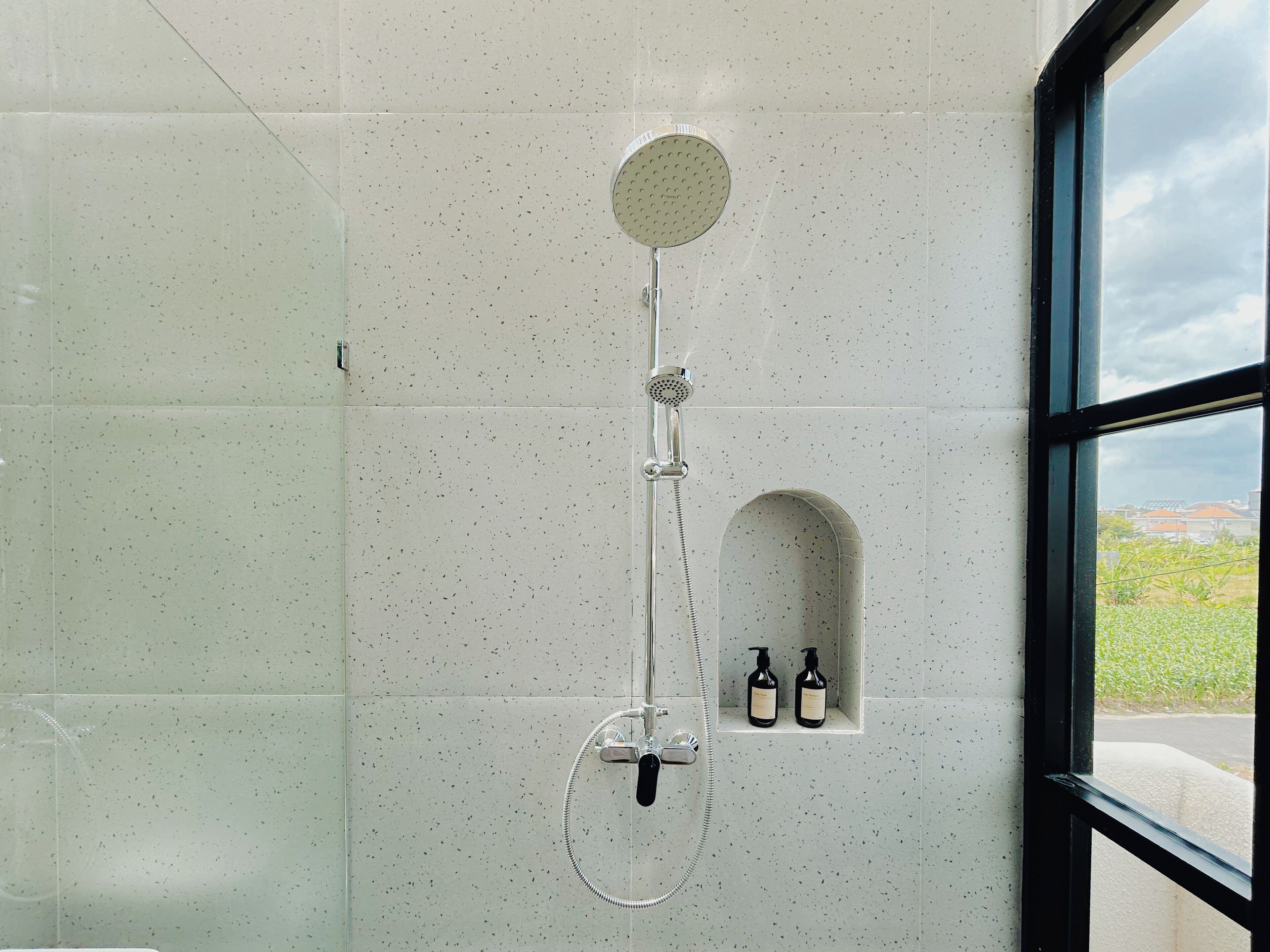 Een douche met een handdouche en een muurbevestigde kraan, een klein plankje met twee flessen, en een raam met uitzicht op groen en gebouwen.