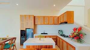 A modern kitchen with wooden cabinets, a central island, and a dining area with wicker chairs. - Beachside Luxury 3BR Mediterranean Sanur (Denpasar)