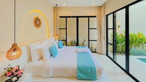 A modern bedroom with a large bed, a decorative wall piece, a hanging light fixture, and a view of greenery outside. - Beachside Luxury 3BR Mediterranean Sanur (Denpasar)