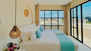 A modern bedroom with a large bed, a hanging light fixture, and a view of the city through large windows. - Beachside Luxury 3BR Mediterranean Sanur (Denpasar)