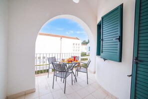 A balcony with a table and chairs, a view of buildings, and a door with green shutters.