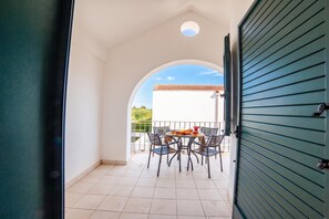Una puerta abierta que da a un balcón con mesa y sillas, vista de un edificio con tejado de baldosas y cielo despejado.