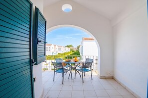 Un balcón con una mesa y sillas, vista a edificios y una puerta con contraventanas.