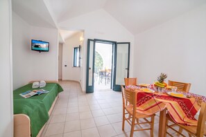 Une pièce comprenant un lit, une table à manger avec des chaises, un téléviseur et une vue sur l’extérieur par une porte ouverte.