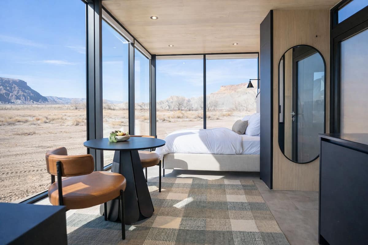 A modern room with a bed, a dining table, and chairs. It has large windows offering a view of a desert landscape.