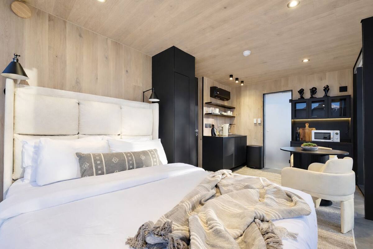 A modern bedroom with a large bed, a bedside table, a chair, a desk, and a kitchenette.