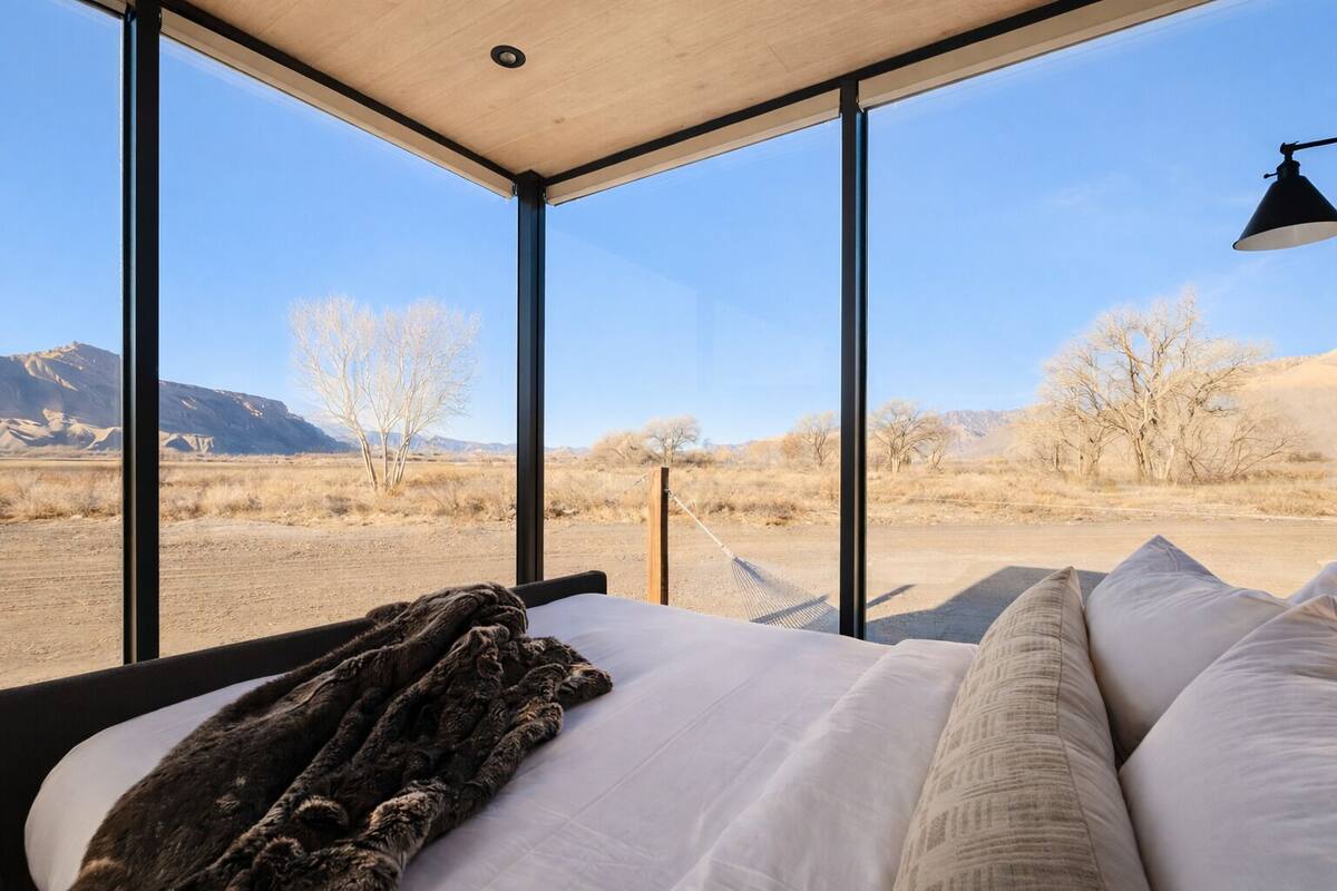 A bedroom with a large window, a bed with white linens, a dark throw blanket, and a black lamp.