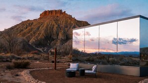 A modern building with a large reflective glass facade, a seating area with a bench and a trash can, and a view of a mountainous landscape. - Reflections (Torrey)