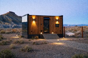 A modern wooden cabin with a glass door, two exterior lights, and a hammock.