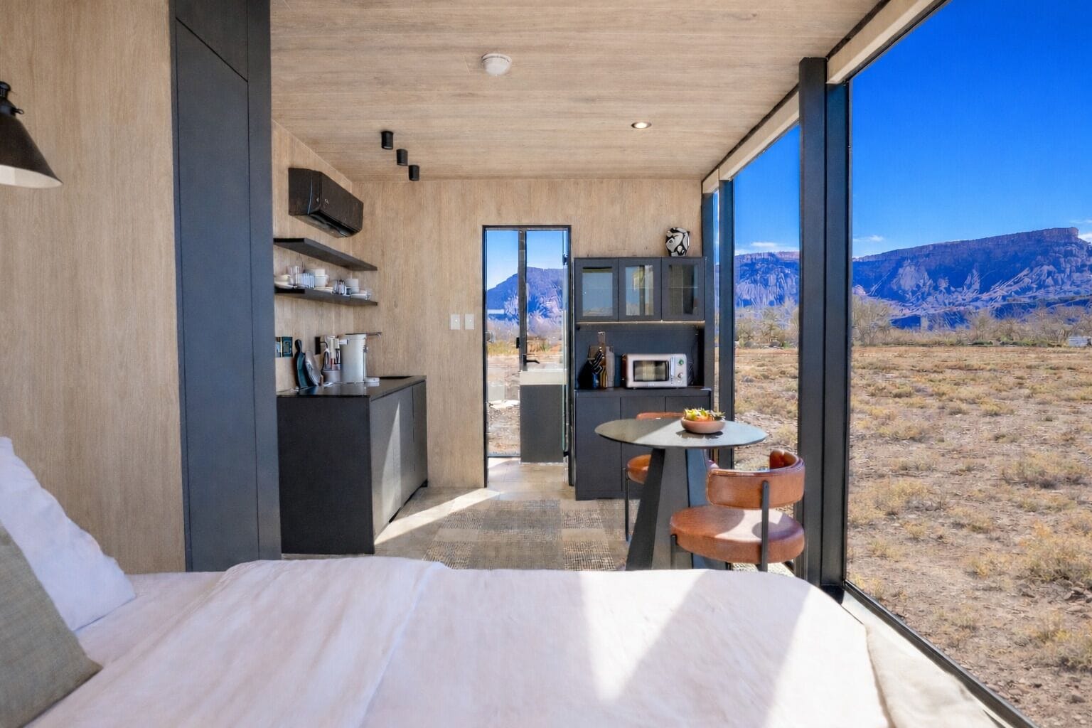 A bedroom with a large bed, a small table with chairs, a kitchenette, and a view of the mountains.