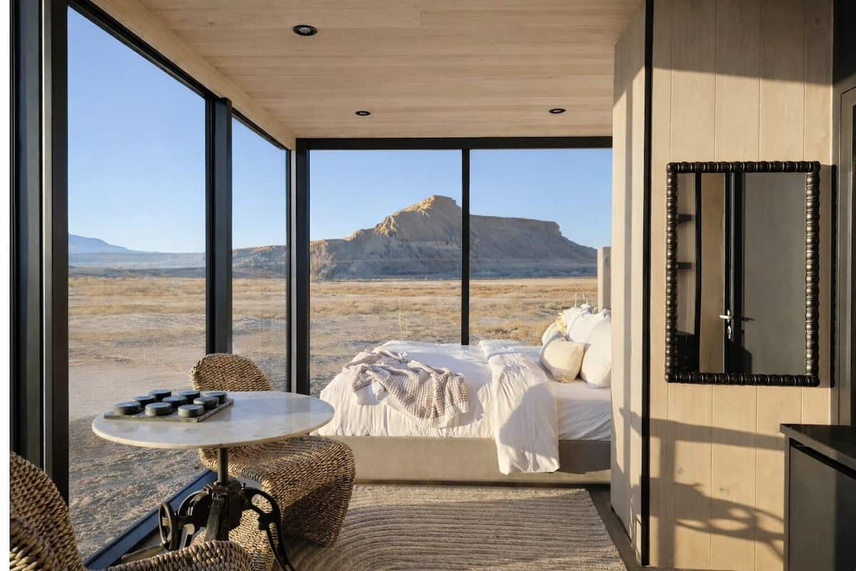 A bedroom with a large window, a bed, a round table, and a woven chair.