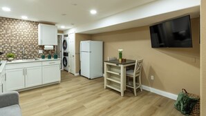 Une cuisine compacte avec des meubles blancs, un lave-linge encastré, un réfrigérateur et un téléviseur à écran plat fixé au mur.