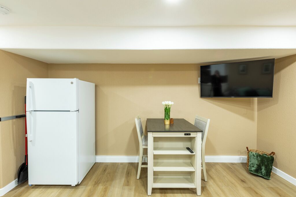 A compact room with a white refrigerator, a table with chairs, a flat-screen TV mounted on the wall, and a small green bag on the floor.