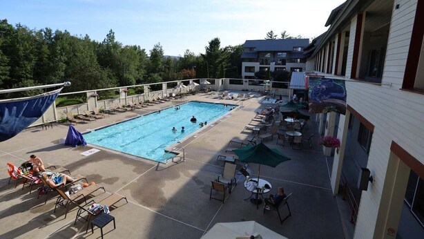 Un espace piscine aménagé avec des chaises longues, des parasols et un bâtiment doté d’un balcon donnant sur la piscine.