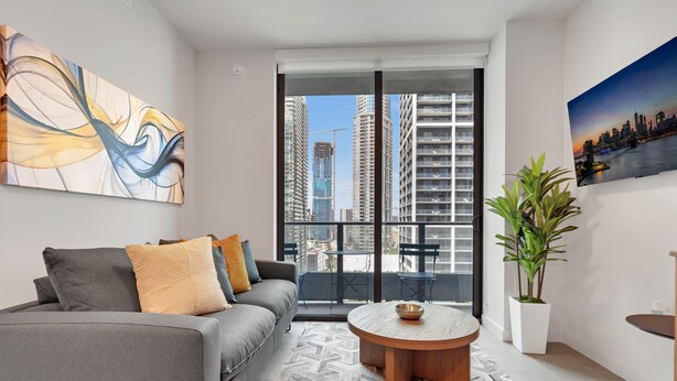 A modern living room with a grey sofa, a round coffee table, a large abstract painting, a potted plant, and a view of a cityscape through a large window. - 501 First Luxury Ocean View from Balcony Rooftop Pool & GYM 2110 (Miami)