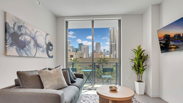 A modern living room with a grey sofa, a round coffee table, a cityscape view through large sliding doors, and a framed abstract painting. - 501 First Luxury 1 BR + Den & 2 Baths Rooftop Pool & GYM 502 (Miami)