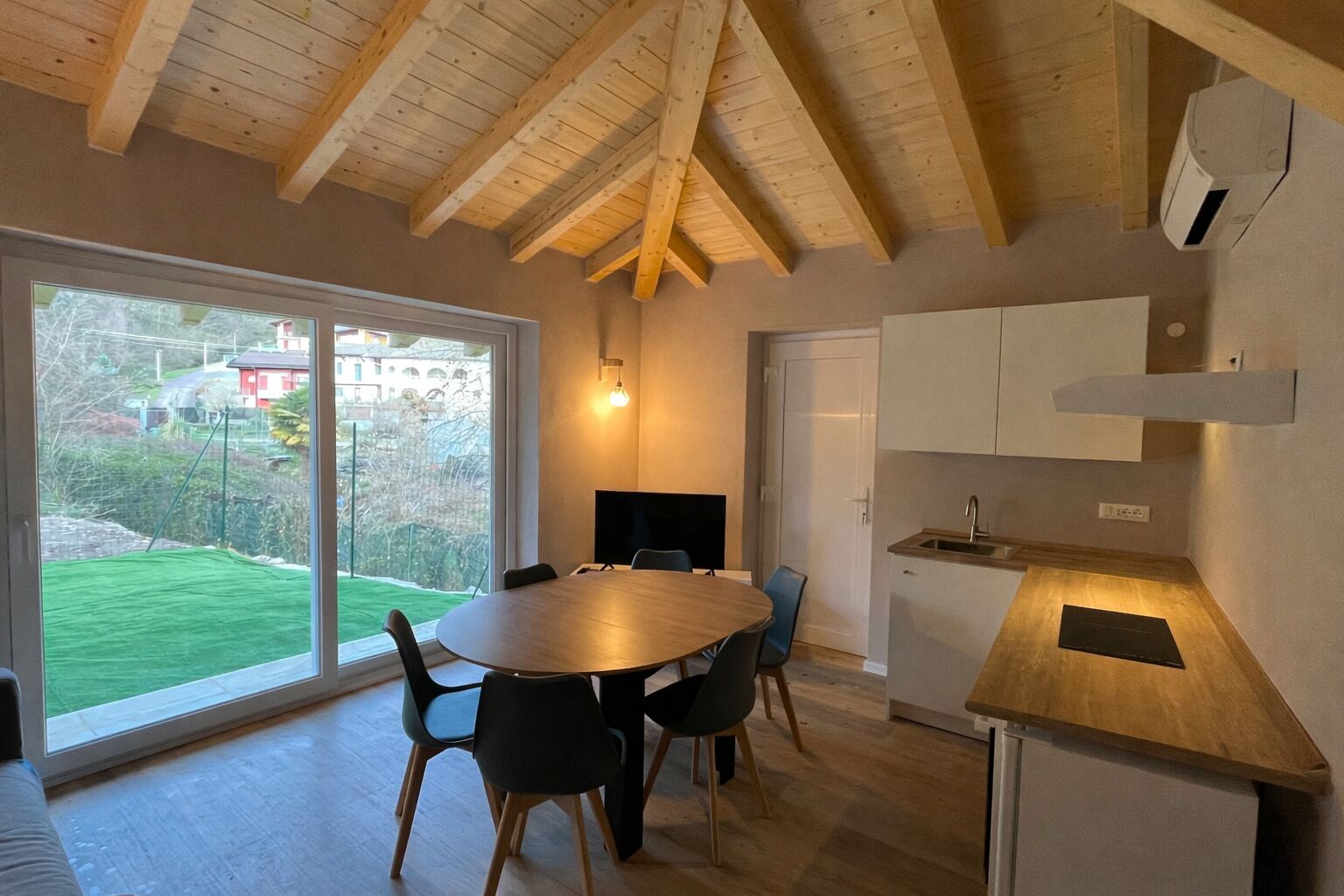 A modern kitchen with a wooden ceiling, a dining table with chairs, a built-in oven, and a large window offering a view of the outdoors.