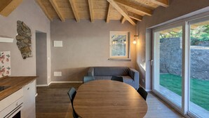 Uma área de refeições com mesa e cadeiras de madeira, parede de pedra e vista para um gramado verde através de grandes portas de vidro deslizantes.