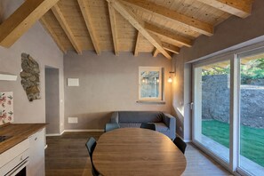 Ein Essbereich mit einem Holztisch und Stühlen, einer Steinwand und Blick auf eine grüne Rasenfläche durch große Schiebeglastüren.